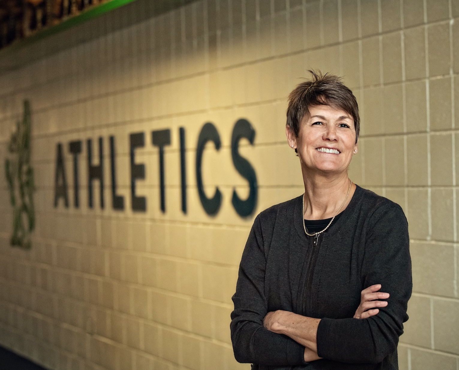 Meg Seng smiling, standing, arms folded, in front of the "Green Hills Athletics" sign.